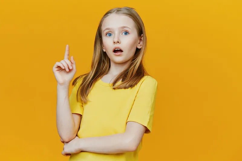 Surprised girl with long hair wearing a yellow shirt against a bright yellow background, expressing curiosity and wonder, ideal for educational concepts