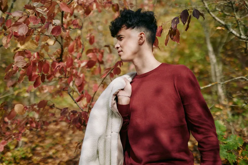 A young man stands in a autumn forest, wearing a burgundy sweater and draped jacket, conveying authenticity and credibility through a candid, natural pose.