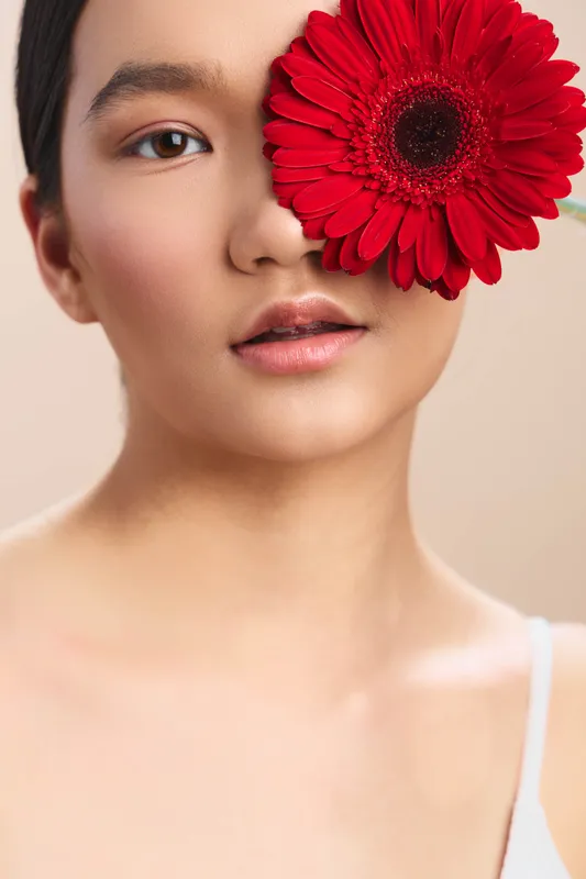 Asian woman presents a close up portrait with a bold red flower partially covering one eye, soft lighting, smooth skin, calm expression, contemporary beauty photography style.