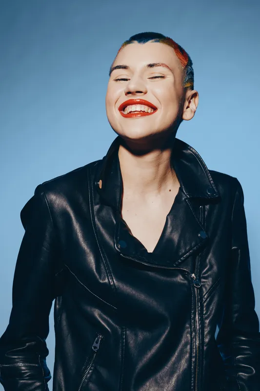 Young woman with short colorful hair and bright red lipstick smiling in a black leather jacket against a blue background, expressing joy and confidence.