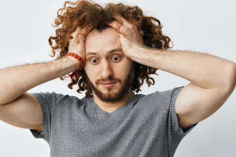 Frustrated young man with curly hair holding his head, expressing anxiety in a minimalistic studio setting, ideal for themes of stress or mental health