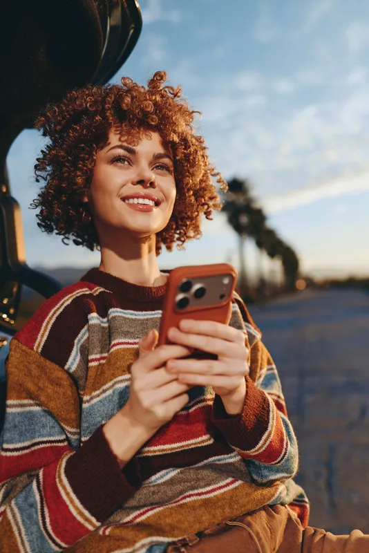 Woman lifestyle portrait of curly hair young woman smiling while holding smartphone outdoors on street at sunset golden hour casual knitwear sweater travel mood relaxed city vibe