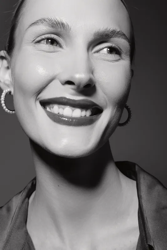 Close-up portrait of a smiling Caucasian woman with light skin and shiny lips wearing hoop earrings in black and white. Concept for beauty, happiness, and natural skincare.
