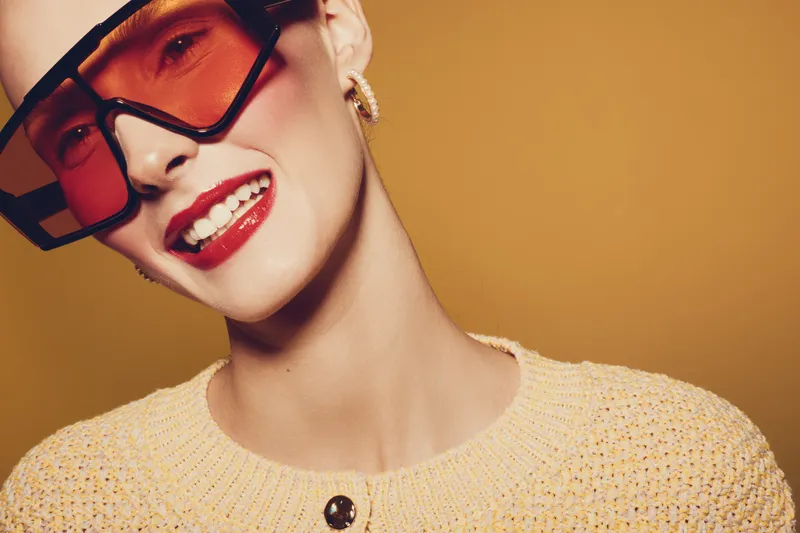 Smiling woman with red lipstick and oversized sunglasses poses against orange background. Fashion concept with trendy accessories and joyful expression in studio lighting.