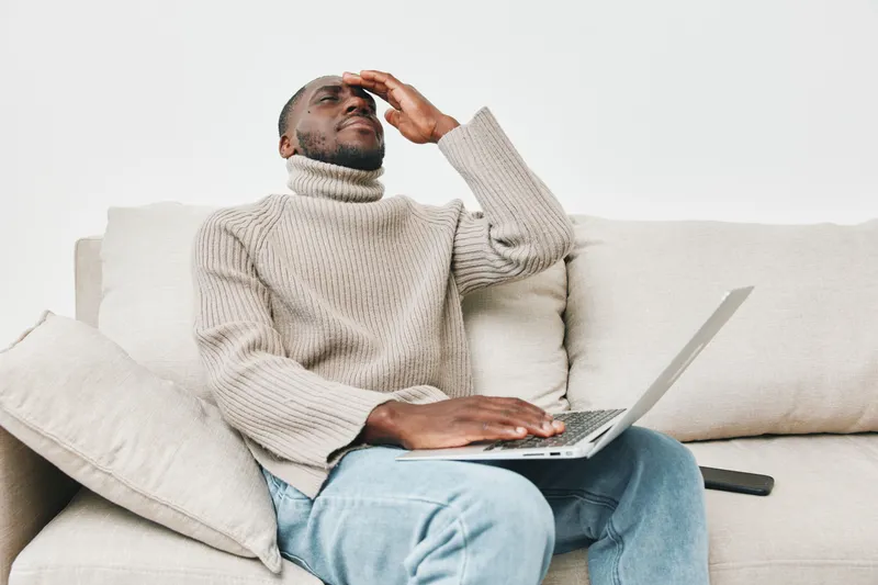 A stressed African man in a cozy sweater sitting on a sofa with a laptop, showcasing emotions of frustration and concentration in a minimalist living room