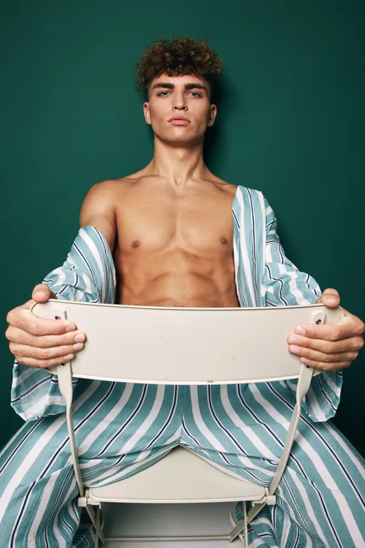 Man with curly hair wearing striped pajamas sitting backward on chair with intense gaze on green background. Shirt open and hands gripping white chair from front.