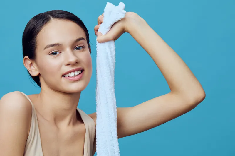 Smiling young woman holding a white towel, showcasing healthy skin against a vibrant blue background, perfect for wellness and skincare themes