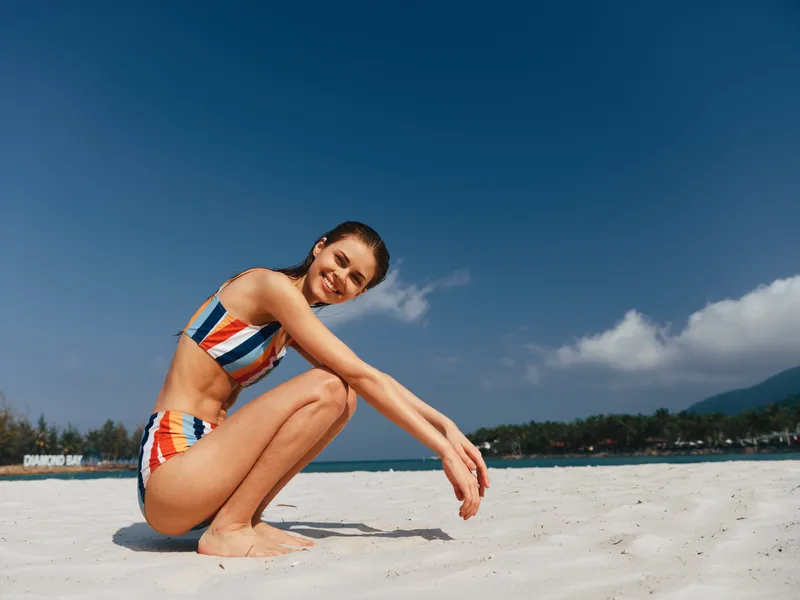 Female model crouches on bright white sand wearing a striped bikini, sunlit figure, relaxed pose, warm tones, clear blue sky, gentle waves, and a vibrant tropical beach mood.