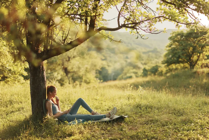 Young woman relaxing under a tree in a green field, enjoying nature and tranquility during a sunny day, evoking feelings of peace and serenity