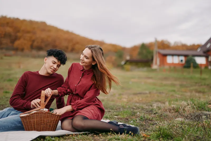 An authentic candid moment of a couple sharing a basket picnic in a sunlit autumn field, relaxed posture, natural smiles, and a genuine connection.