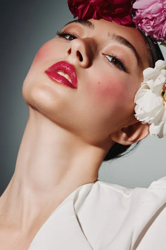 Close-up beauty portrait of a young woman with vivid red lipstick, pink blush, and creative floral headpiece. Fashion and art concept with high contrast lighting.