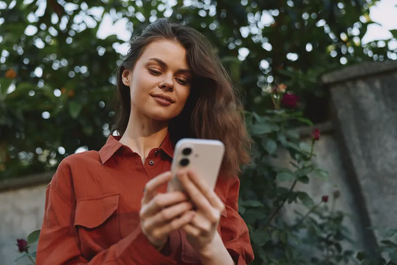 Young woman with long brown hair wearing a rust shirt stands in a leafy outdoor space, focused on her smartphone with a relaxed smile, creating a casual lifestyle moment