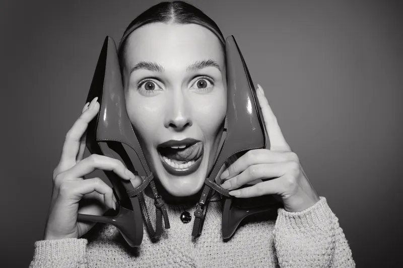 Young Caucasian woman with light skin holds red high heels near her face and makes funny expression. Concept of fashion, style, and playful mood in studio.