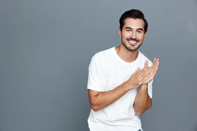 Smiling cheerful young man in a white t-shirt clapping hands, looking at the camera with joy against a plain gray background. People lifestyle concept.