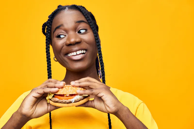 Happy young Black woman enjoying a delicious burger against a vibrant yellow background, showcasing joy and enthusiasm for food, perfect for healthy eating themes
