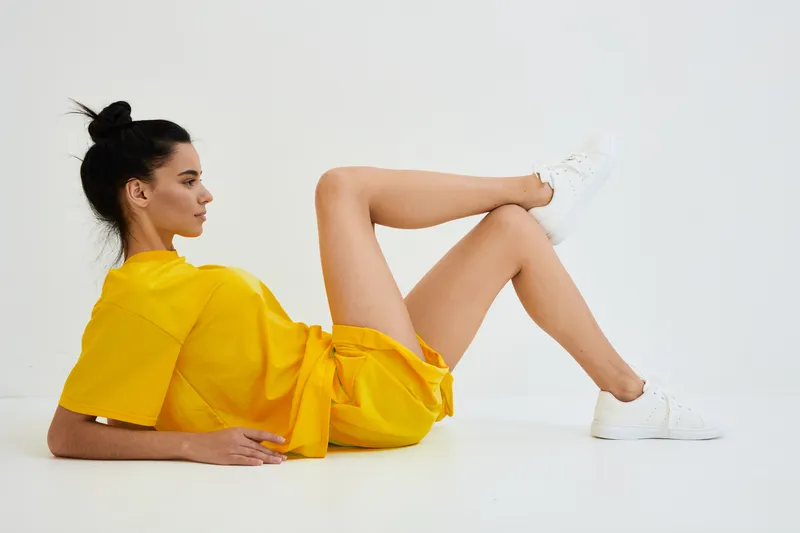 Young woman in yellow outfit lying on white floor, wearing casual sneakers, with a relaxed and confident pose, showcasing minimalistic background and bright fashion style.