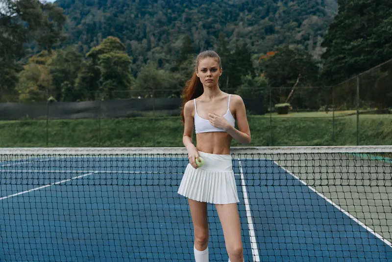 Young female tennis player in white sportswear poses confidently on a tennis court, showcasing athleticism and determination amidst a lush green landscape