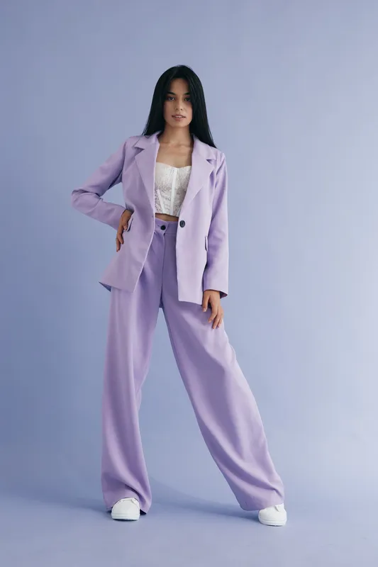 Woman in lilac pantsuit standing confidently against pastel purple background, wearing white sneakers and lace top, displaying modern fashion style and calm expression.