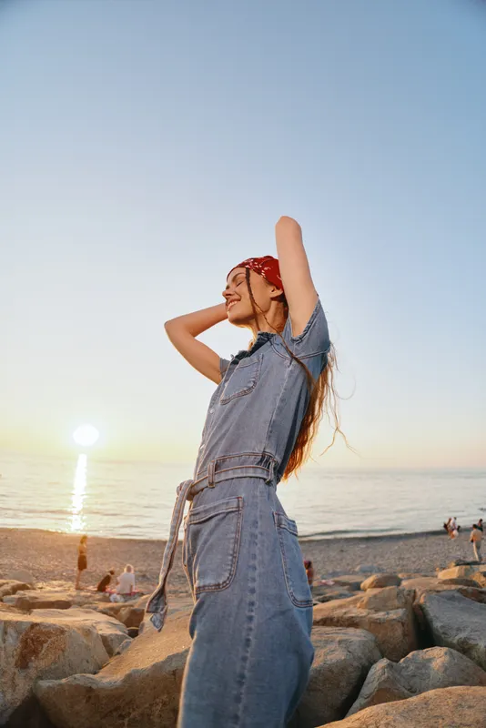 Lifestyle woman in modern boho-western denim street style enjoys sunset by the beach, captured with warm film color tones and relaxed pose.