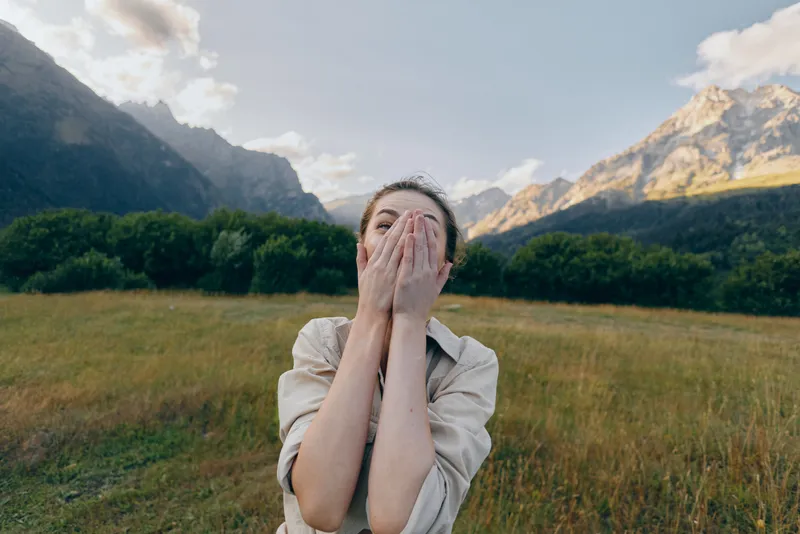 A woman stands in a wide field with towering mountains behind, covering her mouth with her hands in a surprised, candid outdoor lifestyle moment.