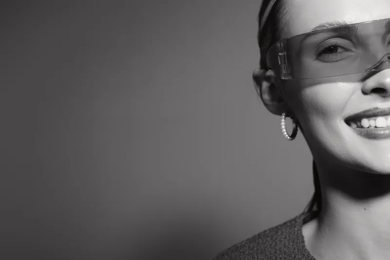 Smiling white woman with fair skin and transparent glasses looks at camera, half face visible. Concept of confidence, fashion, and modern lifestyle in black and white.