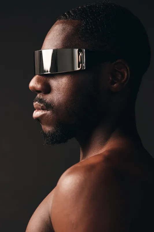 African man with dark skin wearing futuristic silver visor in creative conceptual visual style. Portrait of black male with modern technology accessory on dark background for innovation.