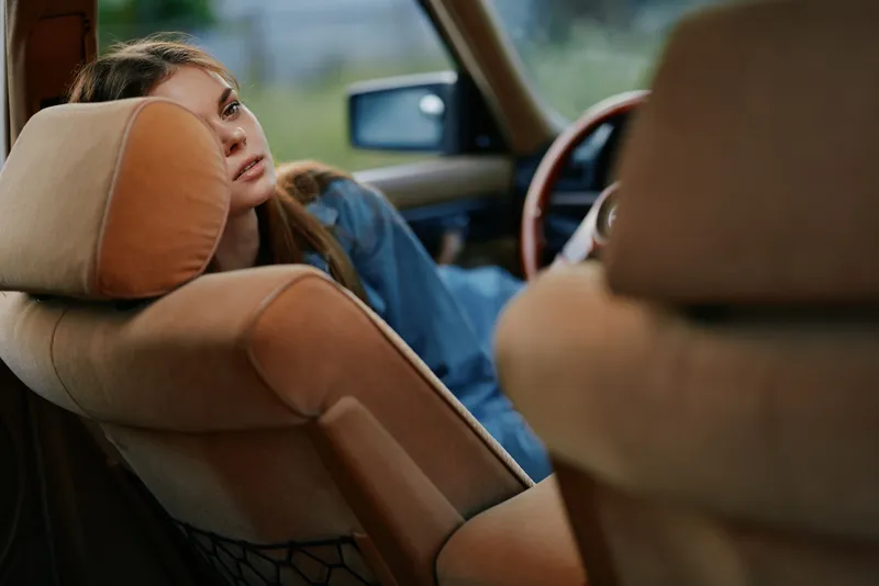 A serene young woman with long hair relaxes in a vintage car, capturing a moment of tranquility in a soft color palette surrounded by nature.