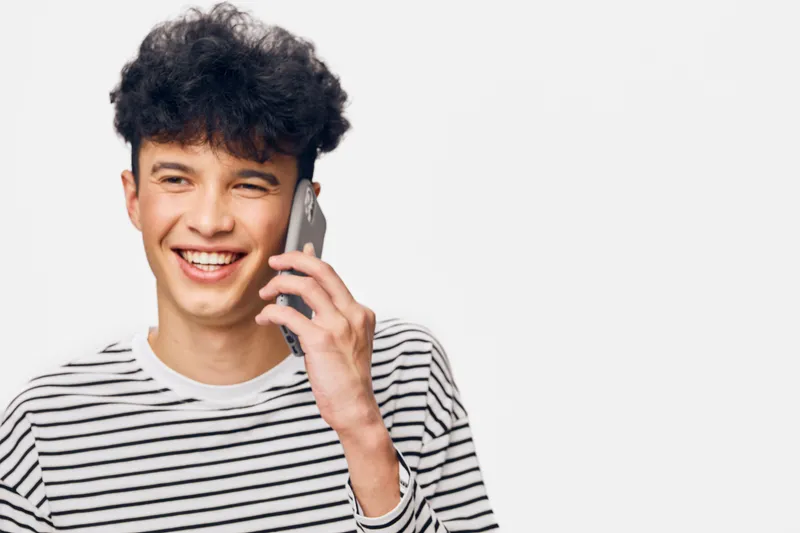 smiling man talking on smartphone wearing striped shirt, cheerful young adult with curly hair against plain white background, casual communication moment