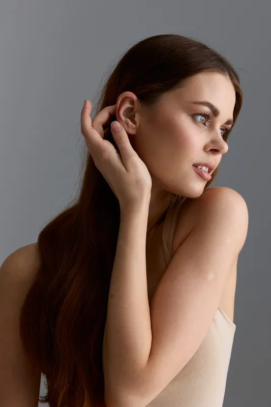 young woman with long brown hair posing gracefully against gray background wearing beige top with natural light highlighting her calm and thoughtful expression