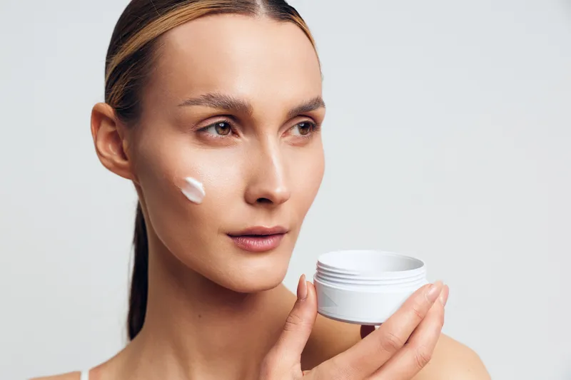 Young woman with clear skin holding white cream jar applying moisturizer on her cheek against a plain background. Concept of skincare, beauty, and daily facial care routine.