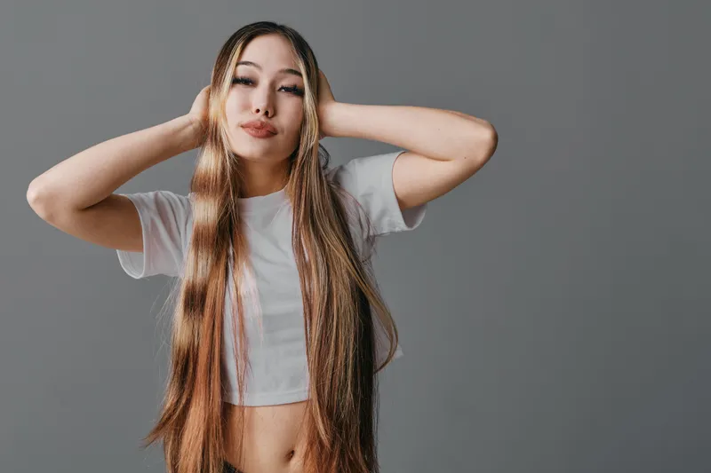 Woman with long braided hair and casual white t shirt posing with hands behind head on gray background. Portrait of young female model with confident expression and stylish look.