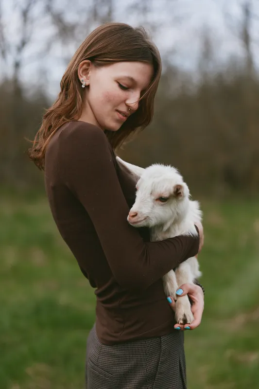 Lifestyle portrait of a woman in a brown long sleeve top embracing a gentle goat kid in a grassy field, calm outdoor mood, soft natural light, intimate rural moment.