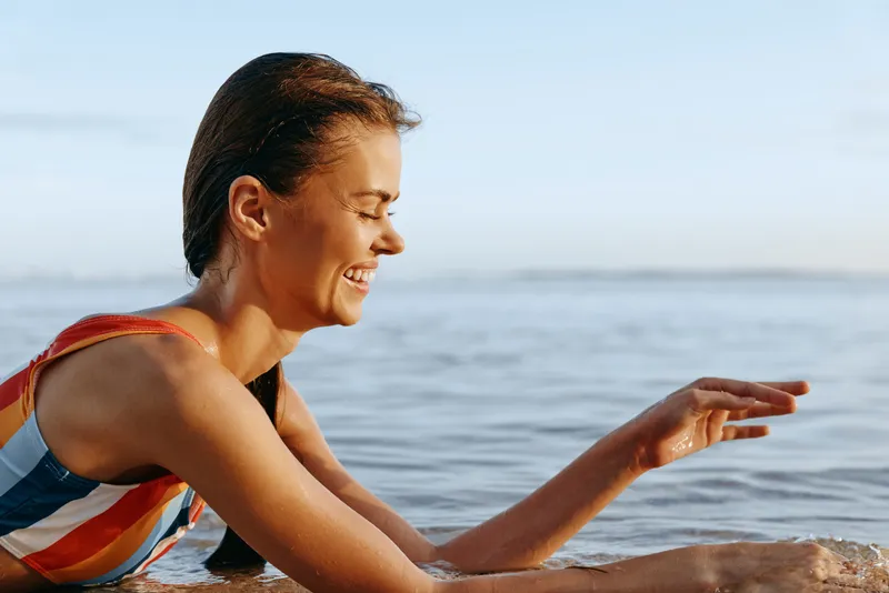Woman applying sunscreen on her arm in the ocean, sea waves in the background, summer beach atmosphere, smiling expression, skincare and self care concept, natural daylight, calm water