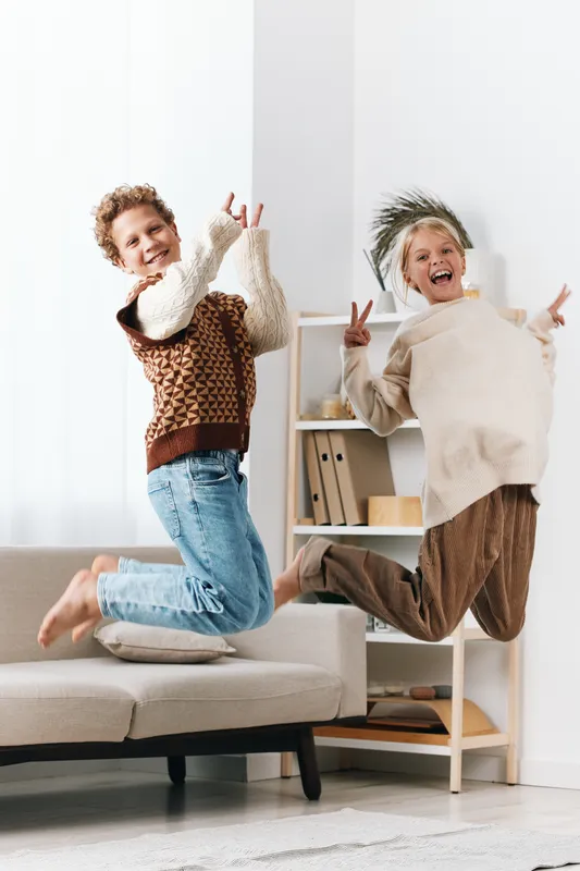 Two children leap with joy on a sofa, mid air, in a bright indoor scene with warm light, capturing a dynamic moment of playful friendship and carefree energy.