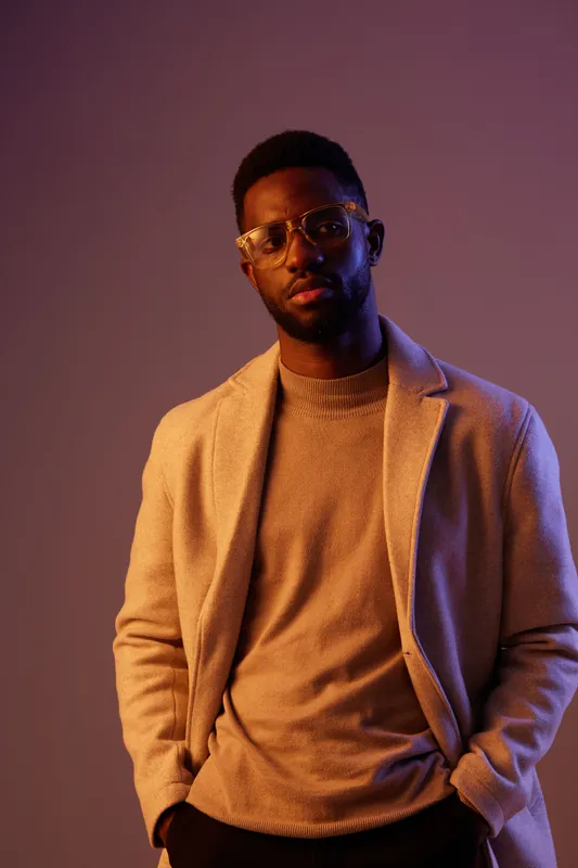 Stylish young Black man in glasses wearing a beige jacket against a colorful gradient background, exuding confidence and modern fashion sense
