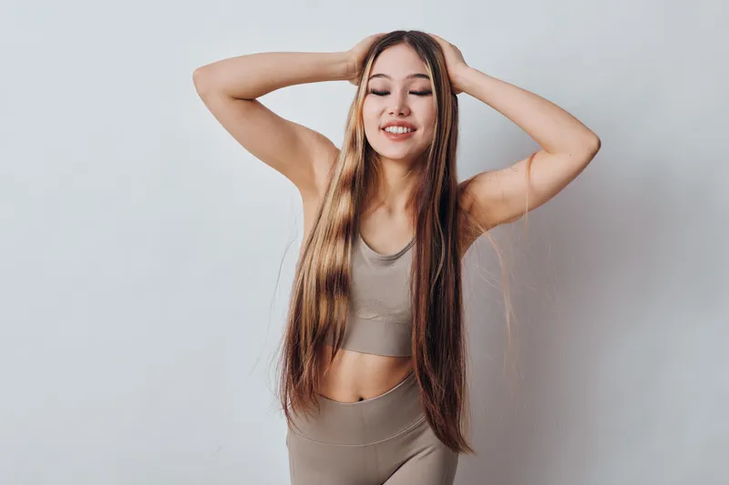 Young woman with long straight hair in beige sportswear posing with hands on head and smiling against plain background. Fitness, beauty and wellness concept with natural look.