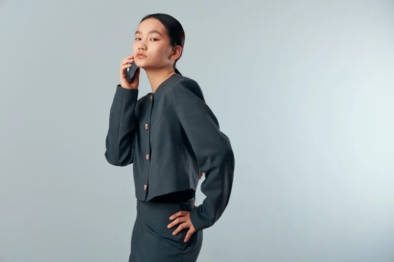 Young woman posing in dark jacket and skirt standing with hand on waist and thoughtful expression isolated on light background for fashion and portrait photography concept.