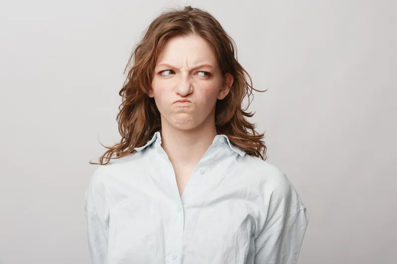 Young woman with curly hair makes a disgusted face, wearing a white shirt against a plain light background. Concept of negative emotions, displeasure, and facial expression.