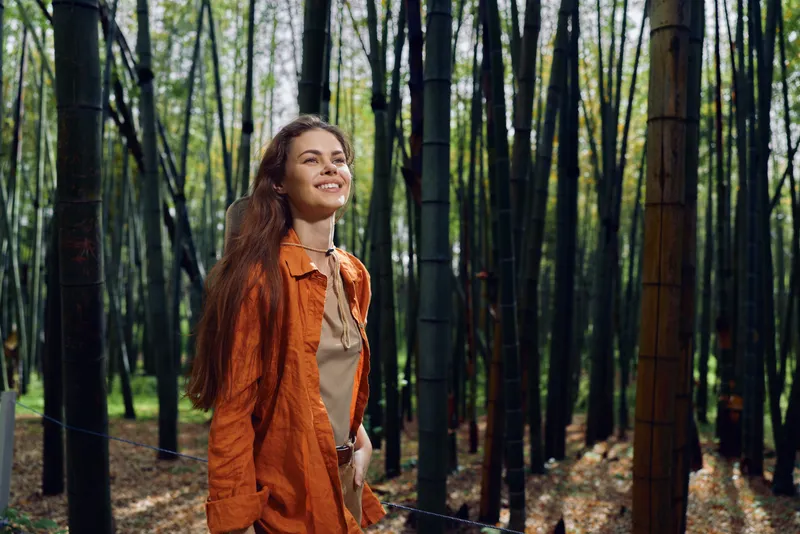 Woman smile bamboo forest nature outdoors portrait happy young adult in casual orange jacket walking among tall bamboo trunks, relaxed expression and sunlit green background.