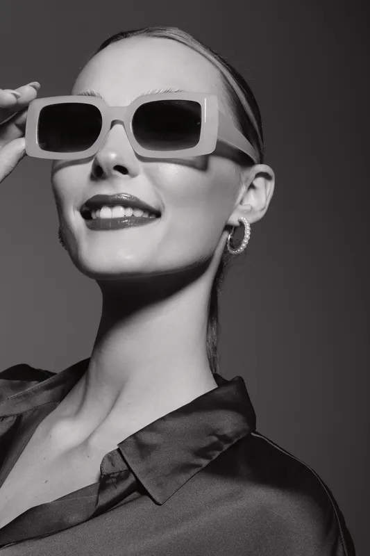 Smiling Caucasian woman with fair skin wearing large sunglasses and hoop earrings poses confidently in a dark blouse. Studio portrait with fashion and style concept.