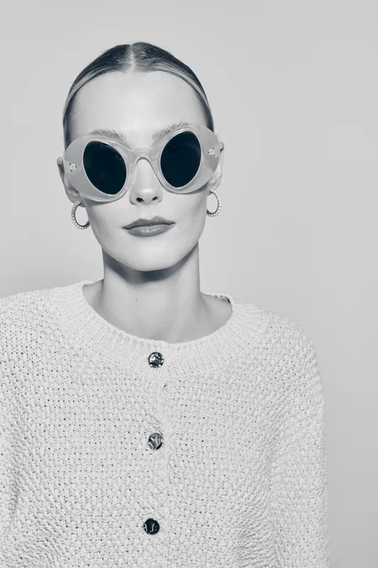 Stylish white woman with pale skin wears large round sunglasses and hoop earrings, posing confidently in knitted sweater in studio setting.