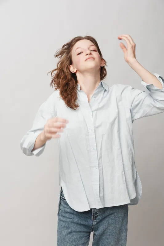 Young woman with curly hair wearing casual shirt and jeans moves freely with eyes closed on light background. Concept of freedom, relaxation, and natural beauty.