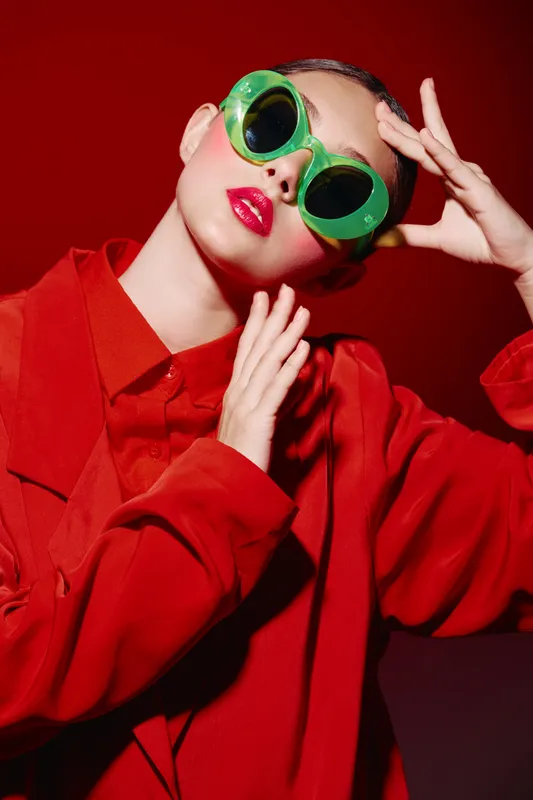 Stylish woman in oversized green sunglasses and vibrant red outfit poses confidently against a deep red background. Concept of beauty, fashion, creative portrait, bold colors.
