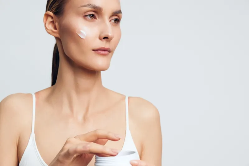Young woman with long brunette hair applies face cream on her cheek while holding an open jar in a white tank top against a plain light background.