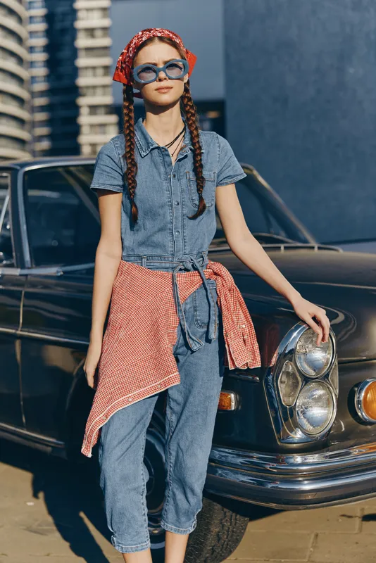 Lifestyle woman in modern boho-western denim street style wearing sunglasses and a red headscarf, standing confidently by a vintage car in warm film color tones.