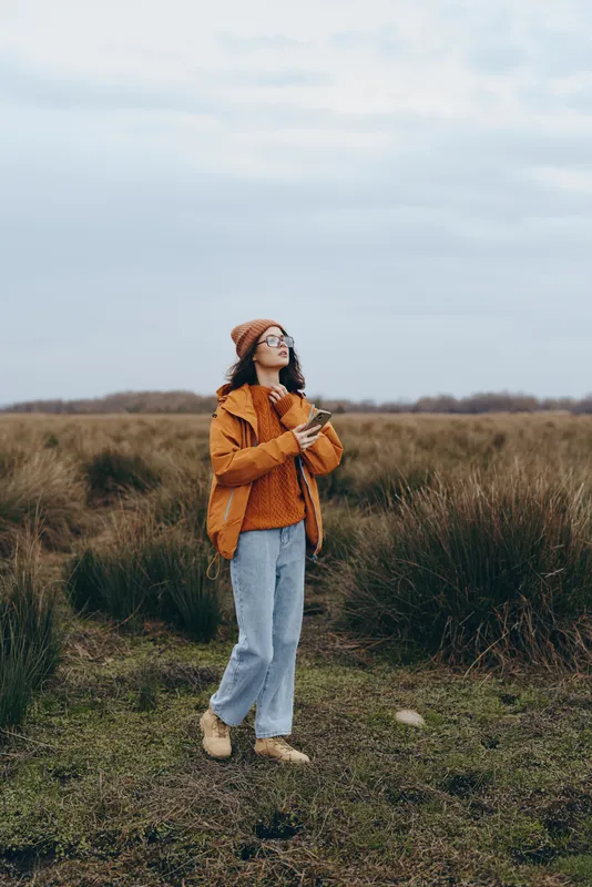 Woman outdoors lifestyle in a grassy field, walking on a dirt path, wearing orange jacket and jeans, knitted hat, calm expression, holding scarf, cloudy daylight, muted autumn colors