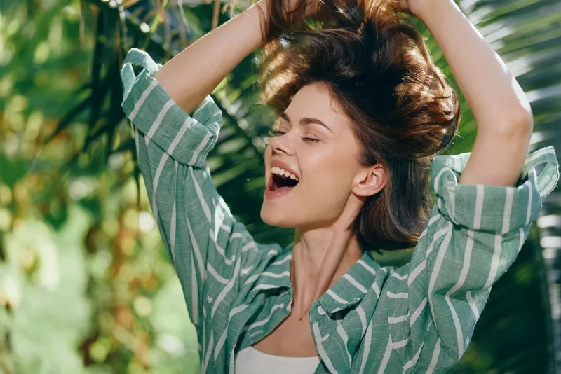 Joyful woman outdoors, hair tossed by wind, laughter and freedom radiate as she moves against lush greenery.