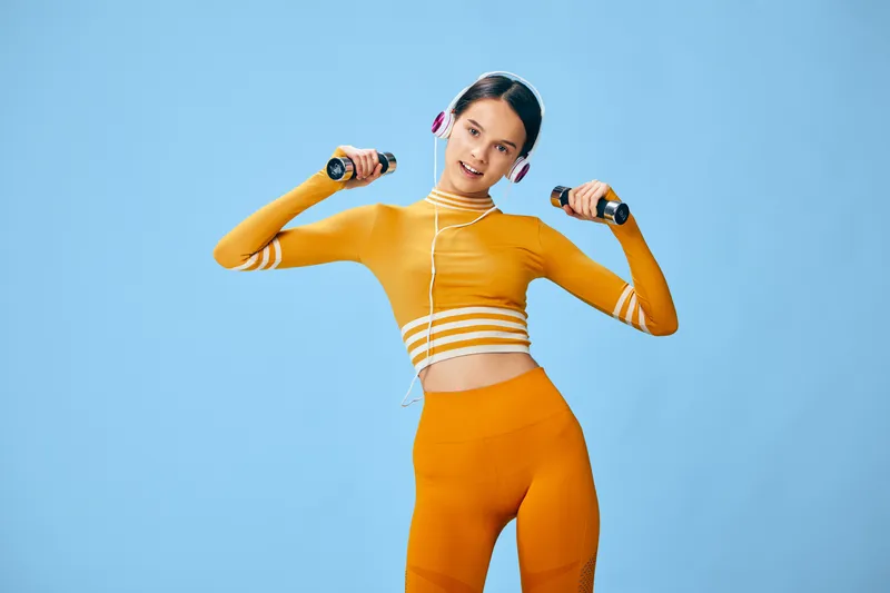 Woman fitness portrait: fit young woman in bright orange activewear lifting small dumbbells while wearing headphones, smiling and exercising against a clean blue studio background, energetic sport