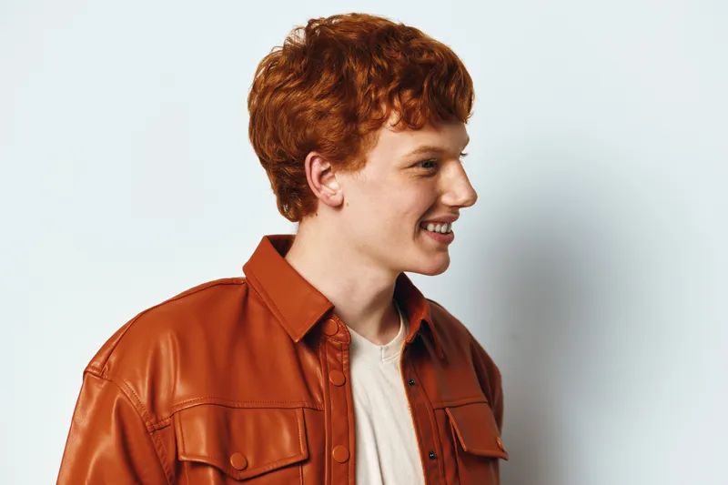 Young man with red hair smiling, wearing brown leather jacket and white shirt on white background, casual style and happy expression in studio portrait session.