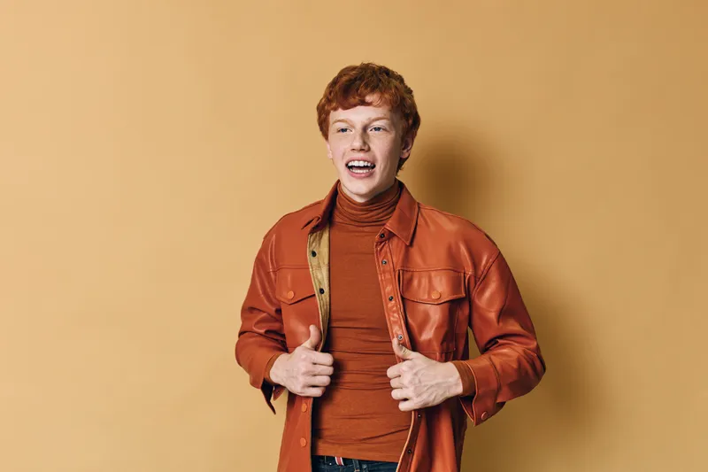 Young man with red hair wearing orange jacket and turtleneck sweater standing on beige background smiling and holding jacket edges for casual fashion style portrait.